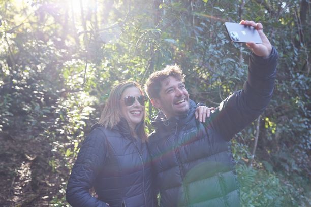 a young couple with puffer jackets smiling with their arms around each other taking a selfie in the rainforest with sunlight shining on them