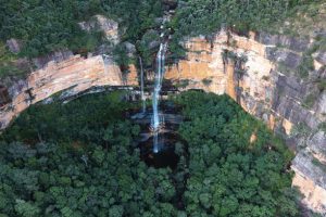 A towering sandstone cliff covered in rainforest with a waterfall cascading down into a circular pool at the bottom.